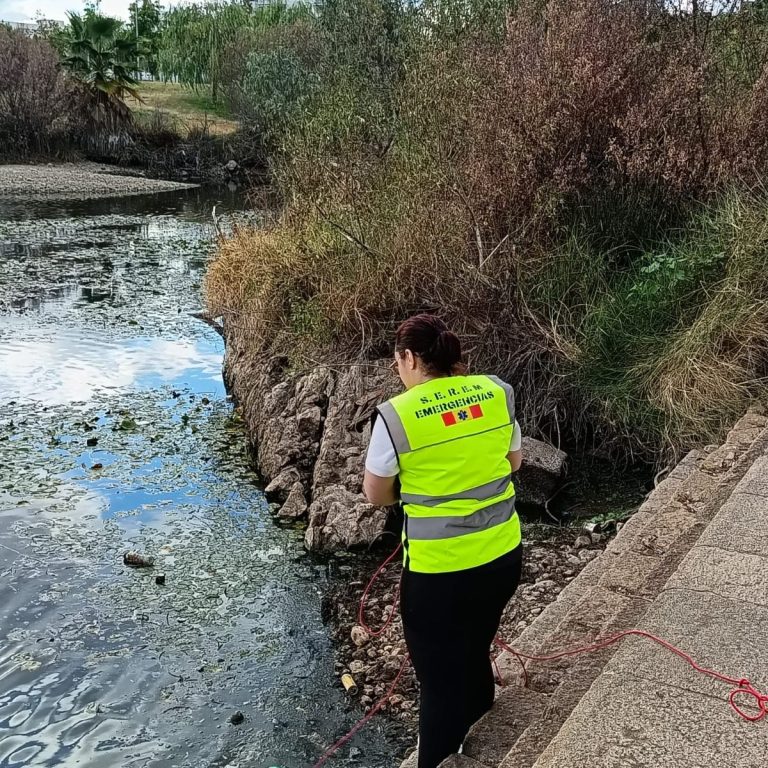 Persona con chaleco reflectante recoge basura cerca de un cuerpo de agua.